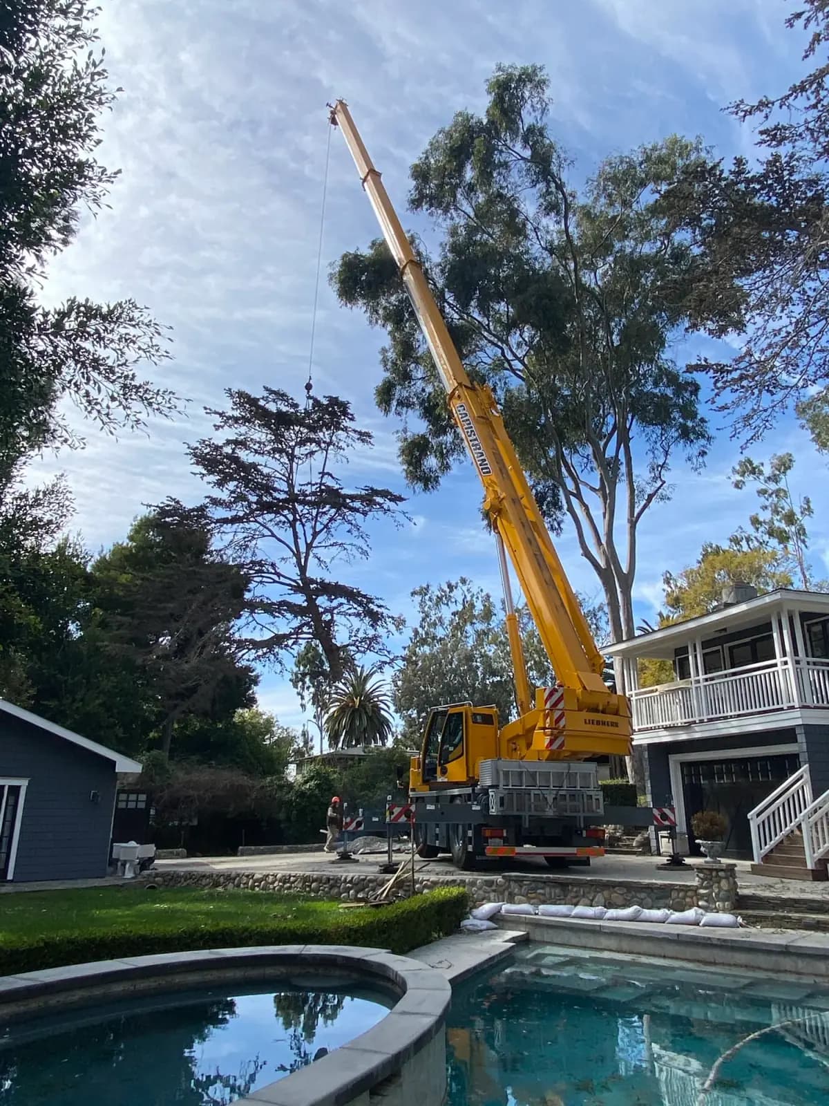 A mobile crane set up on a residential street alongside a tall conifer, outriggers down and boom raised