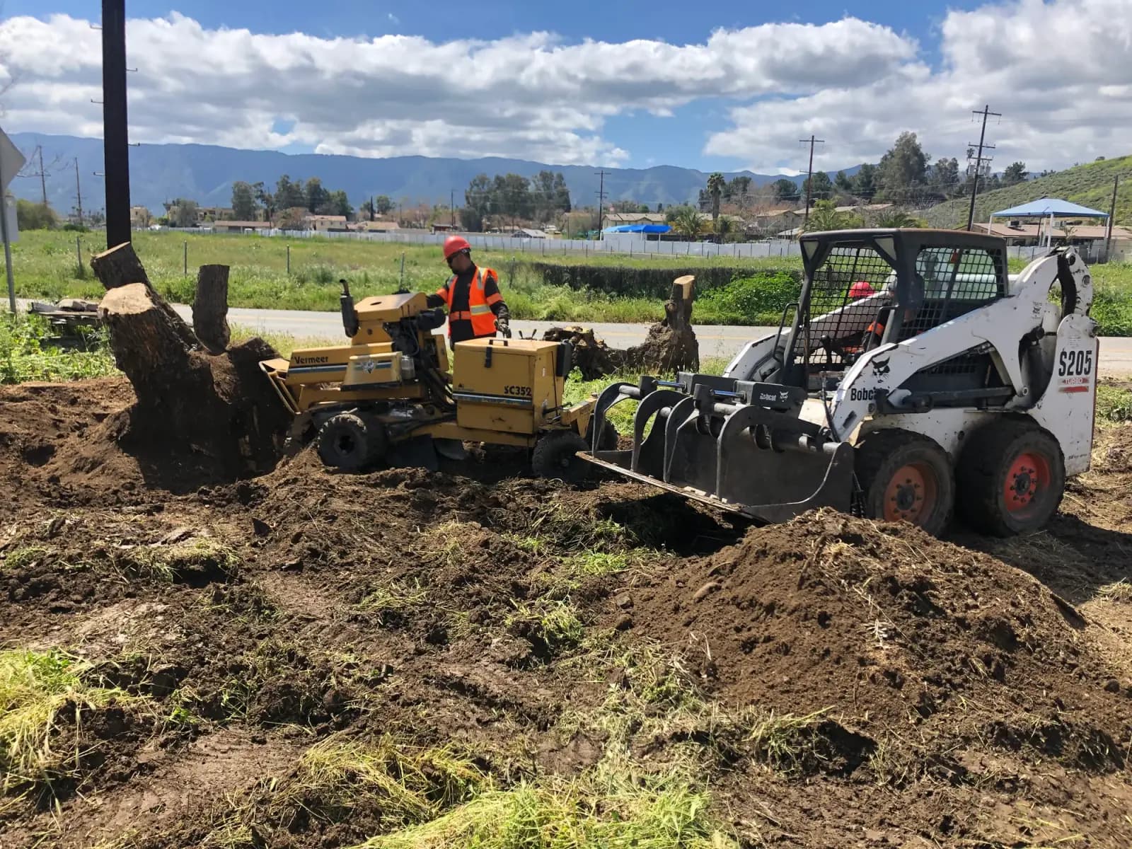 A stump grinder and skid steer clearing an open lot, ground stump debris windrowed across the soil