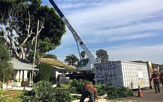 A truck-mounted crane lifting cut sections over a single-storey home during a tree removal, with a debris box on the street