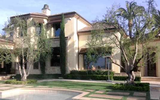 Mature olive trees with open canopies over a lawn inset with cut-stone paving, a stone-coped pool and clipped box hedging in front of a two-storey estate home