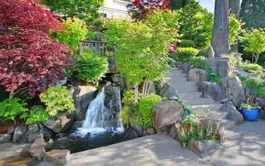 A residential home behind a fully planted front yard, with layered beds, low shrubs and a clipped lawn running to the path