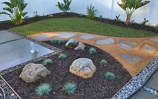 A finished yard with stepping stones set into decomposed granite, mulched beds with boulders and ornamental grasses, and river-rock edging