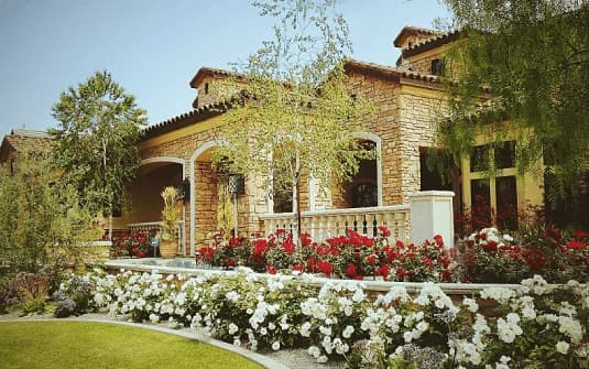 A stone-faced estate home behind a raised planter of massed white and red roses, with mature trees across the frontage