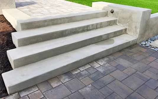 Finished broad concrete steps running between a paver landing above and a paver patio below, with a river-rock margin